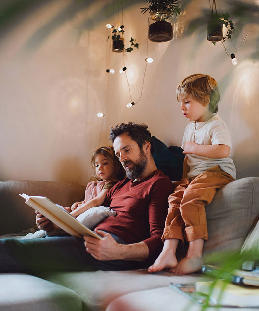 lifestlye-dadwithkids A man sits on a couch reading a book to two young children. Cozy lights and hanging plants decorate the room, creating a warm and inviting atmosphere.