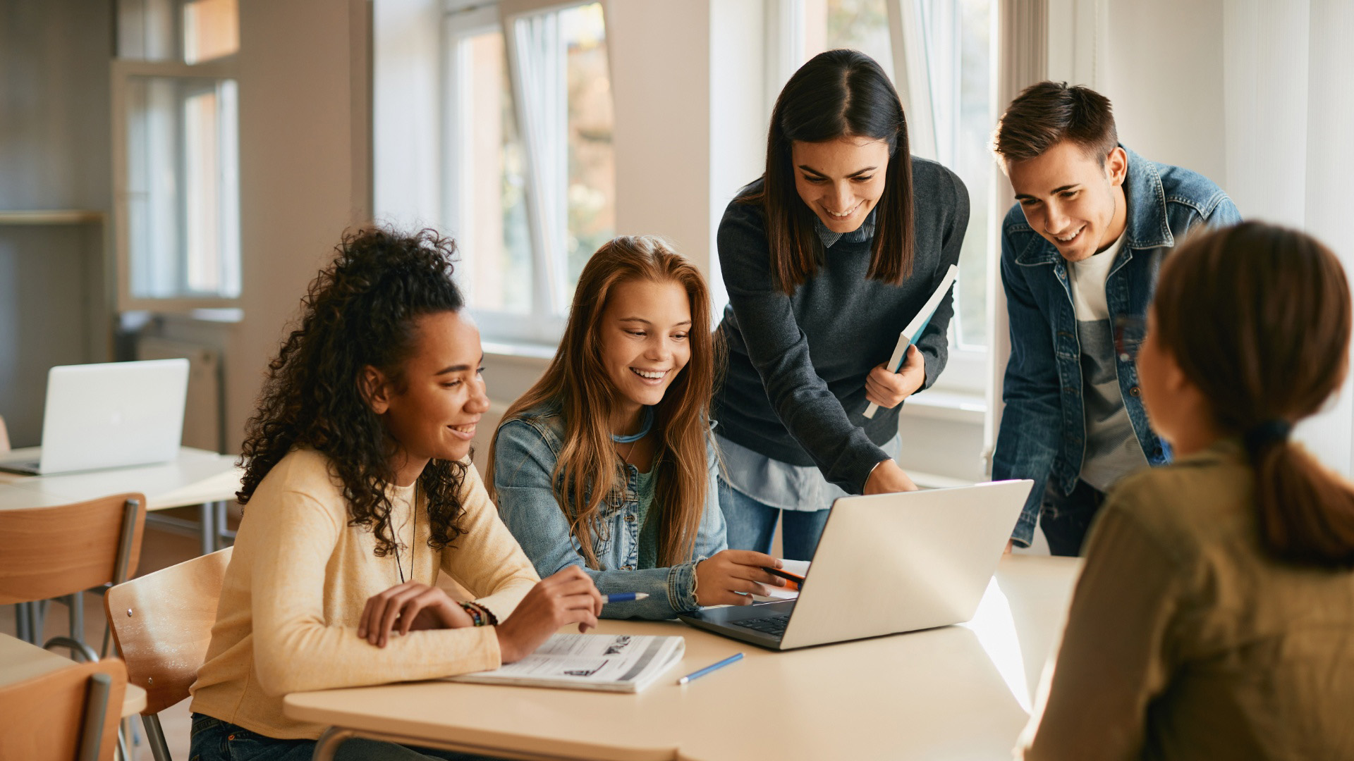 A teacher points to a laptop screen while helping a group of four students from the neighborhood study at a table in a classroom.
