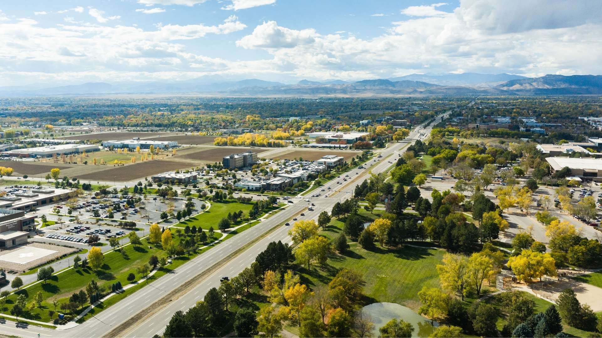 Aerial view of a multi-lane highway running through a suburban landscape with scattered commercial buildings, grassy parks, and autumn trees, with mountains visible on the horizon.