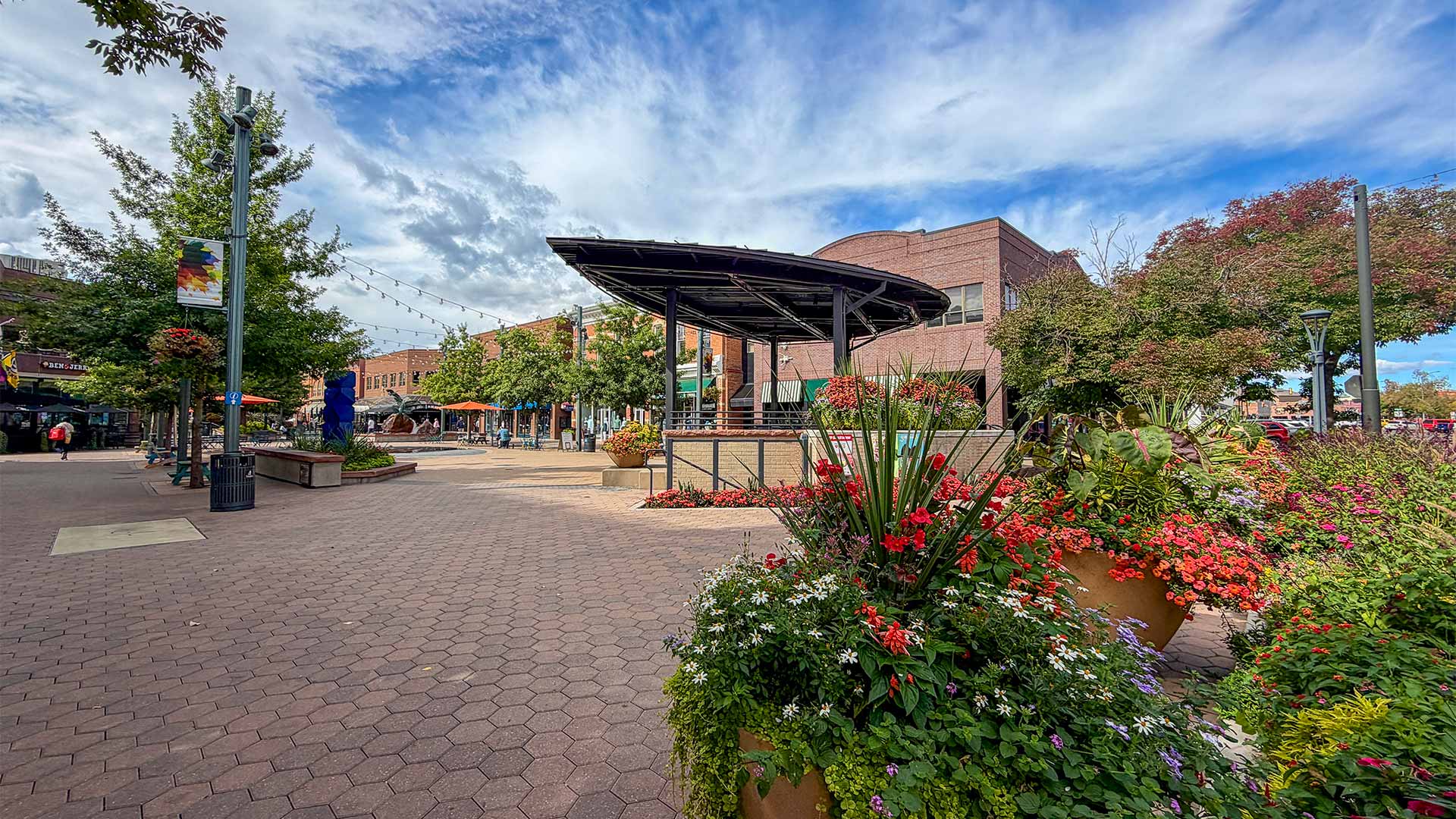 A paved pedestrian plaza with a modern metal bandstand, surrounded by storefronts and large planters overflowing with colorful flowers.