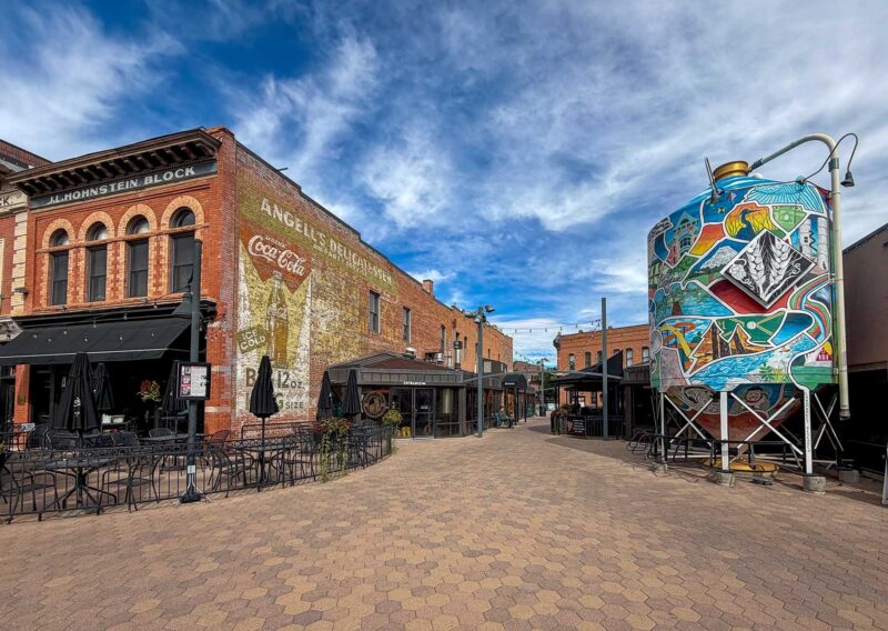 A brick building with a vintage Coca-Cola wall mural stands across from a large, cylindrical art installation painted with a colorful mountain landscape in a pedestrian plaza.