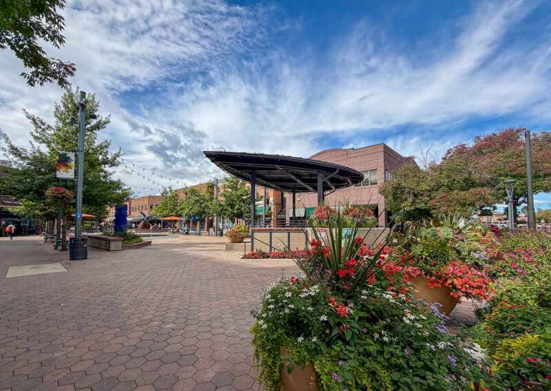 A brick-paved outdoor plaza featuring a large metal pavilion and vibrant flower beds under a blue sky with scattered clouds.